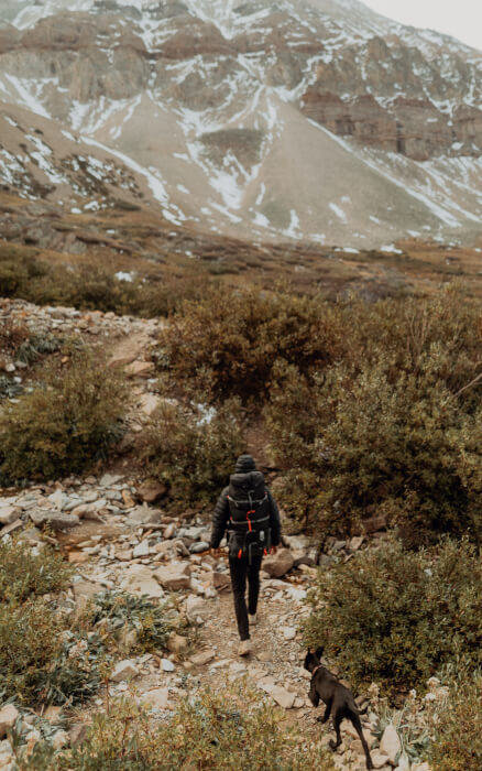 Ein Wanderer geht gefolgt von seinem schwarzen Hund durch eine karge, grasbedeckte Landschaft. Im Hintergrund sind Berge.