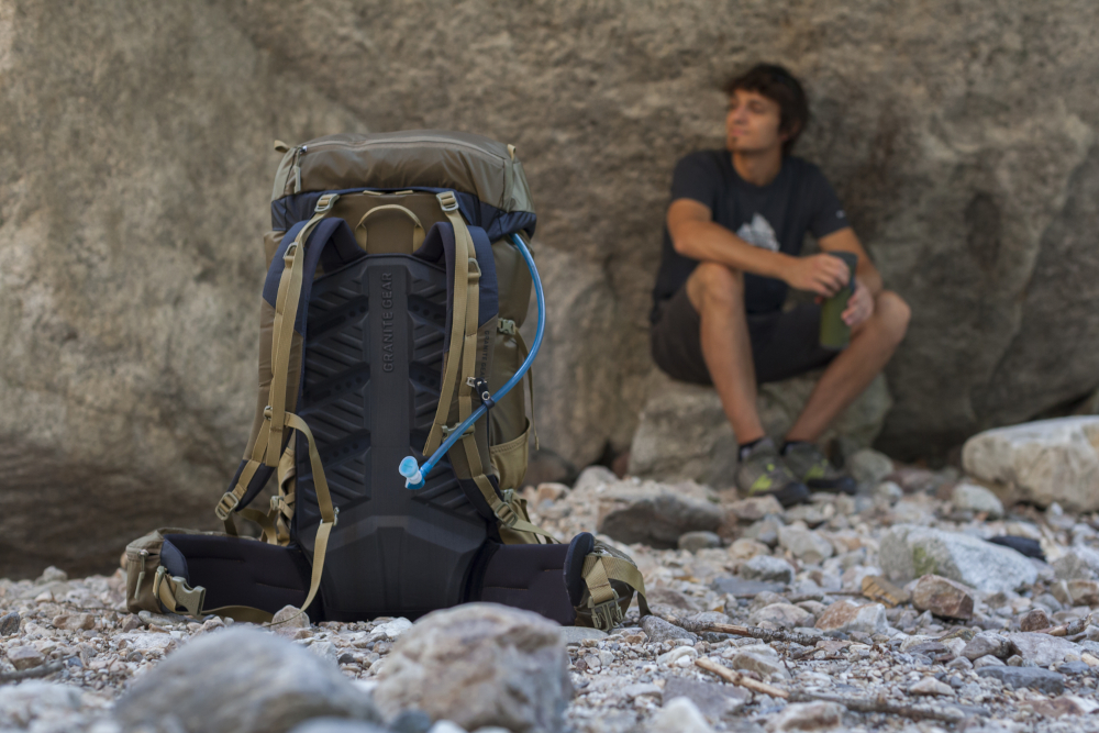 Ein junger mann sitzt an eine Felswand gelehnt. Der Boden besteht aus Geröll. Im Vordergrund ist das Backpanel eines Granite Gear Rucksacks zu sehen.