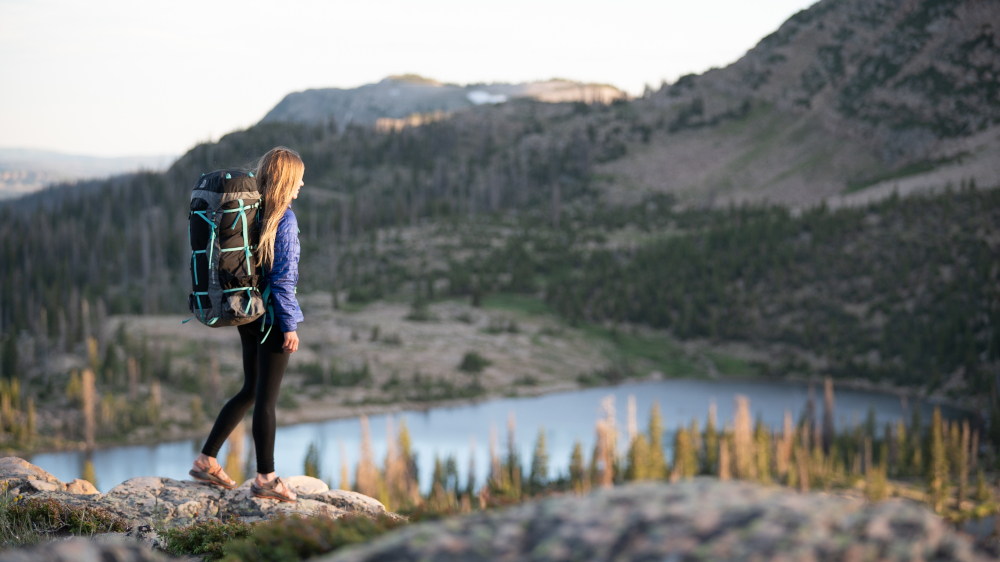 Eine junge Frau läuft entlang einer Felsenkante. Sie trägt einen vollgepackten Rucksack. Im Hintergrund ist ein See und eine Bergkette.