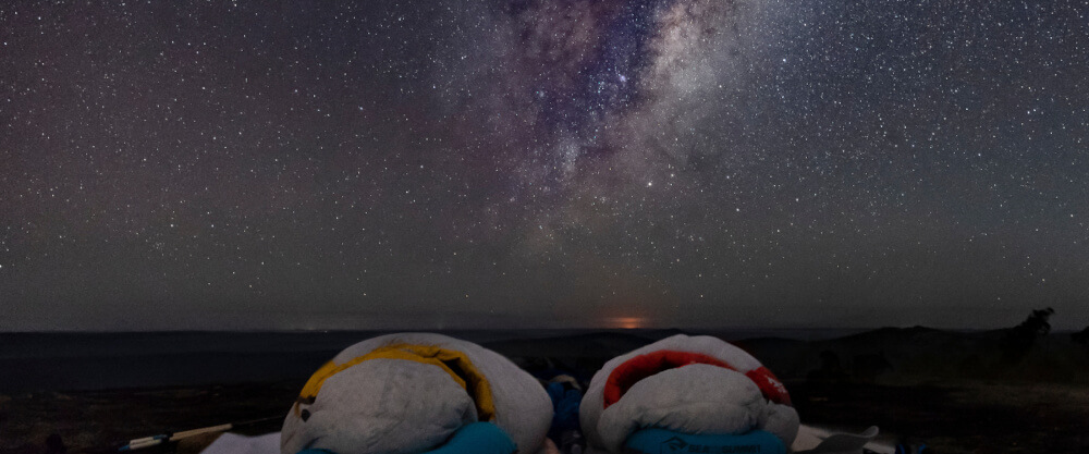Aufmacherbild: Zwei menschen, die in Schlafsäcken unter dem Sternenhimmel liegen