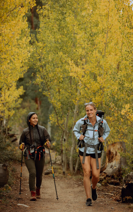 Zwei junge Frauen wandern durch einen gelb gefärbten Laubwald. Beide tragen Rucksäcke