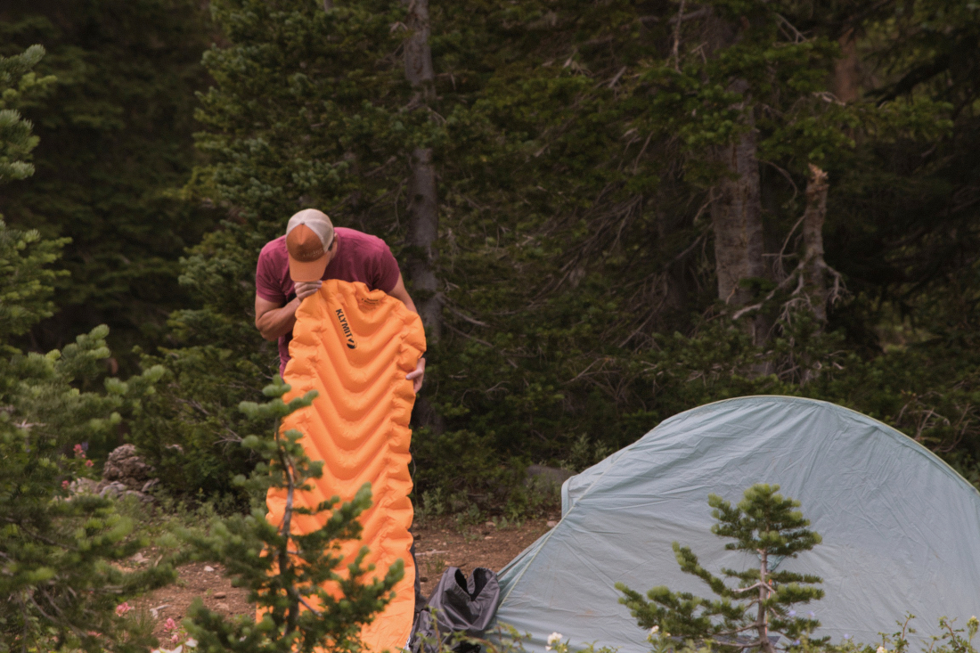 Ein Mann steht im Wald neben seinem Zelt und pustet seine Isomatte auf. Diese ist orange und hat v-förmige Kammern.