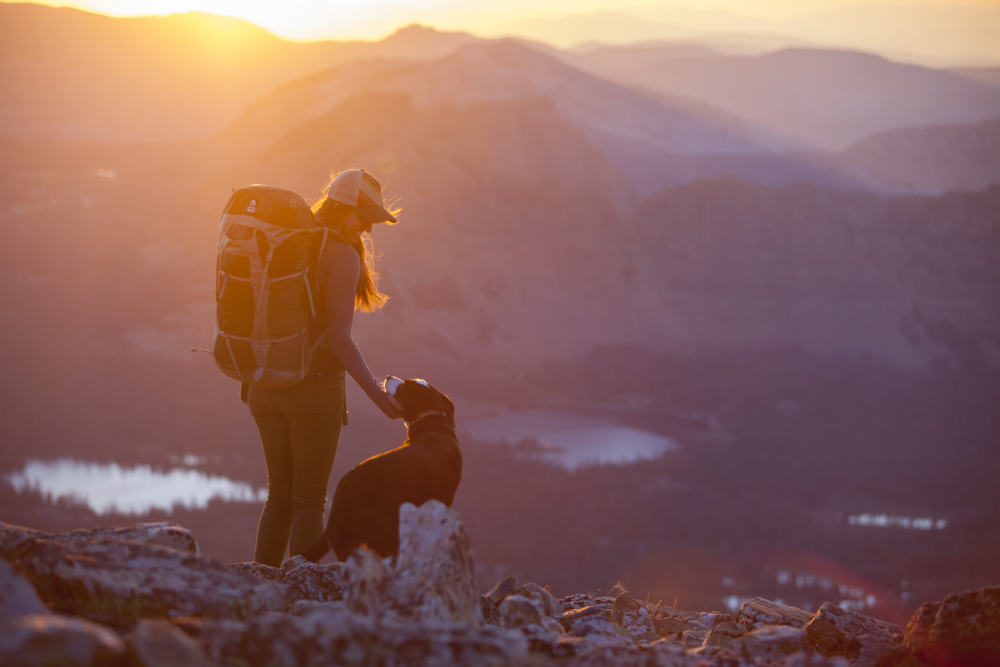 Eine junge Frau mit Cap und Rucksack steht im Sonnenuntergang auf einem Berg. Ihr Blick ist dem hund neben ihr zugewadnt. Im Hintergrund schimmert ein See.
